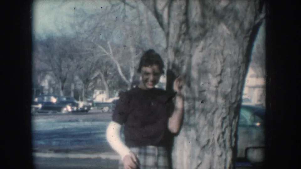 Closer view of woman in dark top and plaid skirt smiling beside a large tree on a 1950s suburban American street, mid-century cars visible in background.