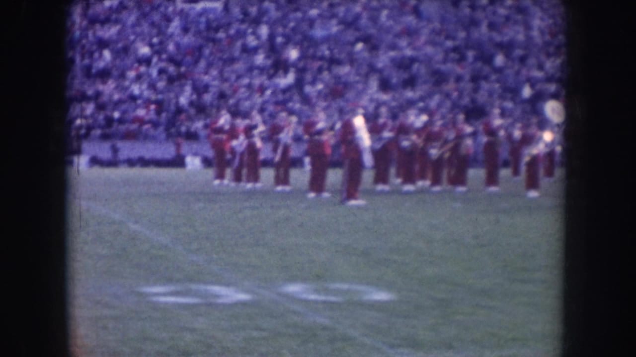 Nebraska marching band in dark uniforms performing in pre-game formation on football field, circa 1957; packed stadium stands visible behind.