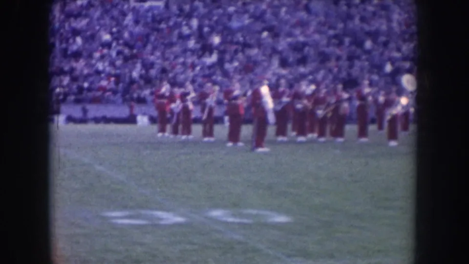 Nebraska marching band in dark uniforms performing in pre-game formation on football field, circa 1957; packed stadium stands visible behind.