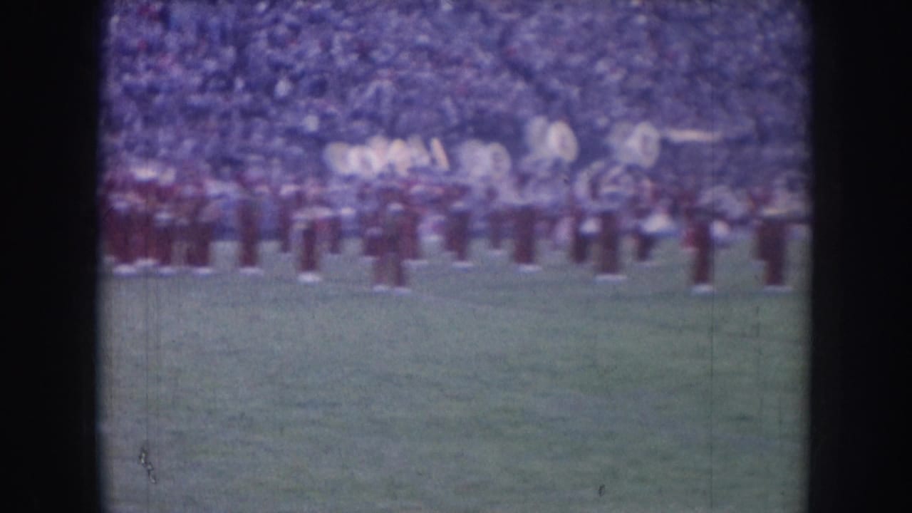Marching band formation on football field with large sousaphones visible at right, stadium crowd fills the stands behind.