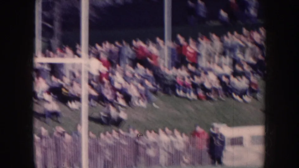 Dense stadium crowd at a Nebraska football game, circa 1957; white goal post structure at left, spectators in mid-century dress packed into the stands.