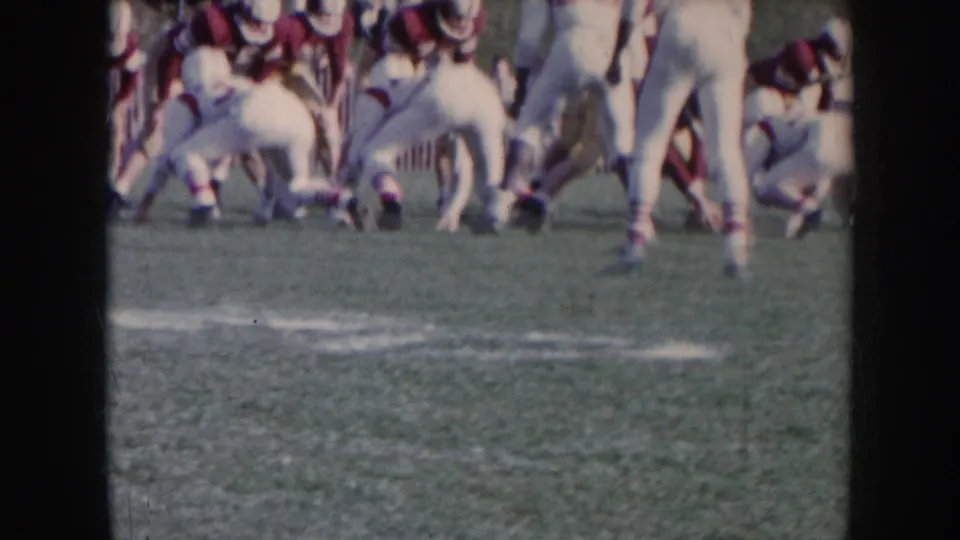Amateur football players in white and maroon uniforms set at the line of scrimmage on a grass field; 8mm footage circa 1957, Nebraska.