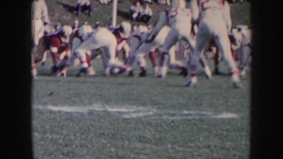 Amateur football players in white and dark uniforms in motion at the line of scrimmage, background field marker and figures visible; 8mm circa 1957 Nebraska.