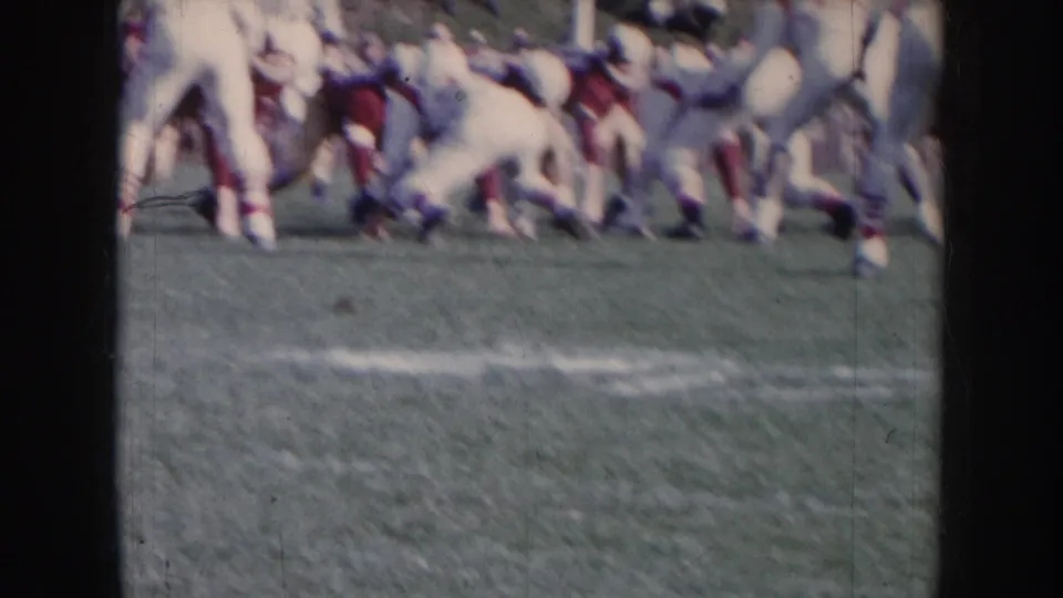 Amateur football players in white and maroon uniforms breaking from the line of scrimmage, motion blur evident; 8mm sideline footage circa 1957 Nebraska.