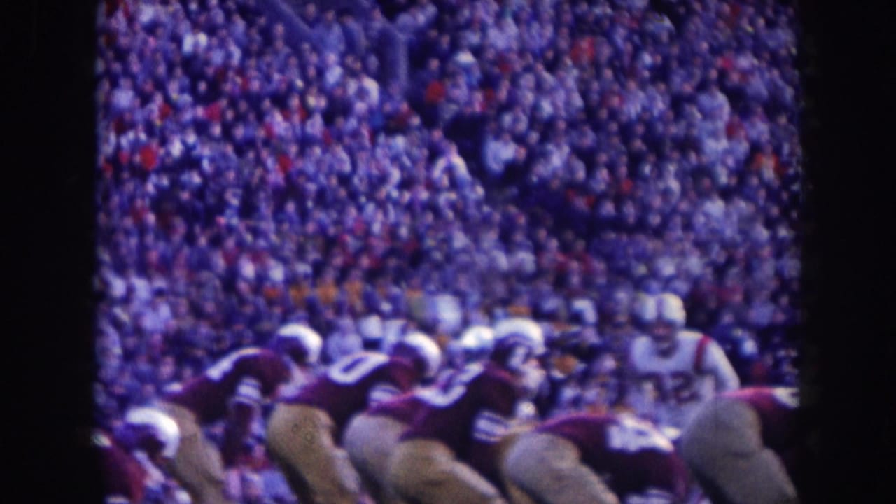 1950s American football players in dark and white uniforms at the line of scrimmage; large stadium crowd behind them; aged 8mm film with purple-blue color cast.