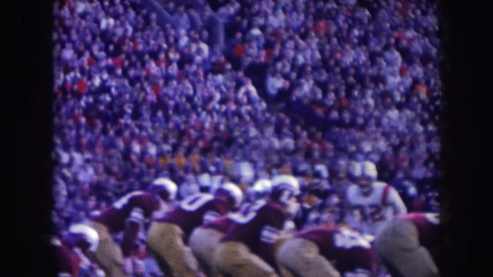 1950s American football players in dark and white uniforms at the line of scrimmage; large stadium crowd behind them; aged 8mm film with purple-blue color cast.