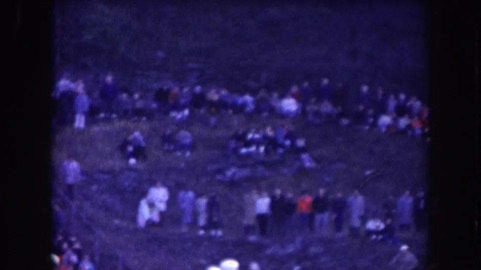 Wide view of what may be the stadium sideline or field perimeter with spectators standing in groups; heavy blue color cast from aged 8mm film stock, circa 1957.
