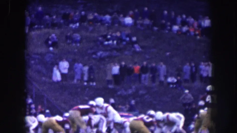 1950s football players in helmets crouching in formation, large crowd visible in packed stadium stands behind them; aged 8mm footage with purple-blue cast, Nebraska circa 1957.