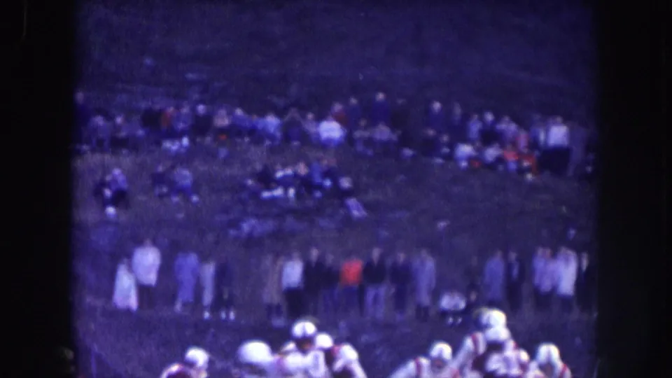 Wide view of a 1950s football field showing players and sideline figures; spectators in period clothing visible in groups; aged 8mm film with blue-purple cast, Nebraska circa 1957.
