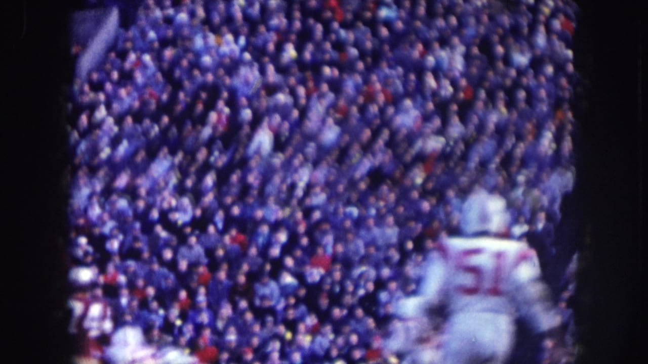 Sideline view of a 1957 Nebraska football game: player no. 51 in white jersey, blue-shifted 8mm color, dense stadium crowd filling the stands behind.