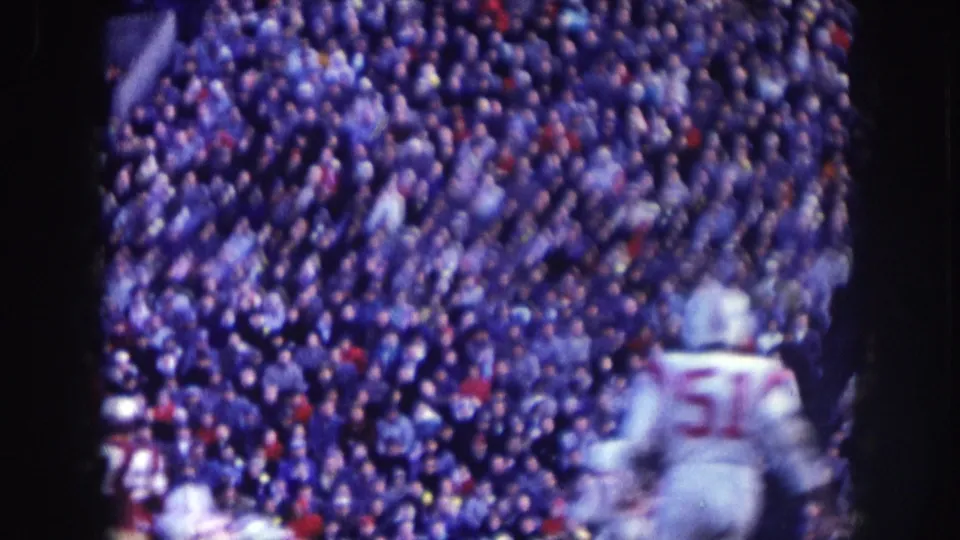 Sideline view of a 1957 Nebraska football game: player no. 51 in white jersey, blue-shifted 8mm color, dense stadium crowd filling the stands behind.
