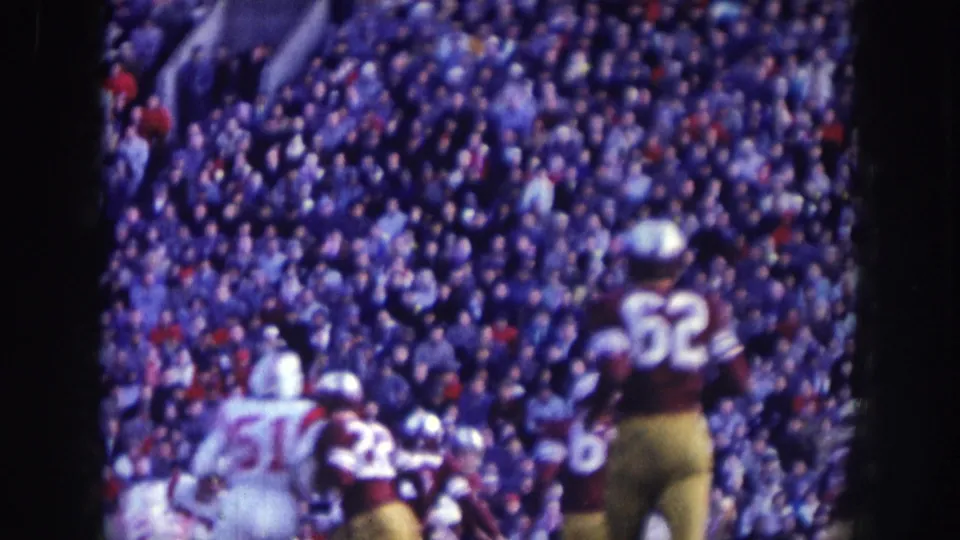 Sideline shot at a 1957 Nebraska football game: player no. 62 in white jersey and an opposing dark-uniformed player, blue-shifted 8mm stock, densely filled stands behind.