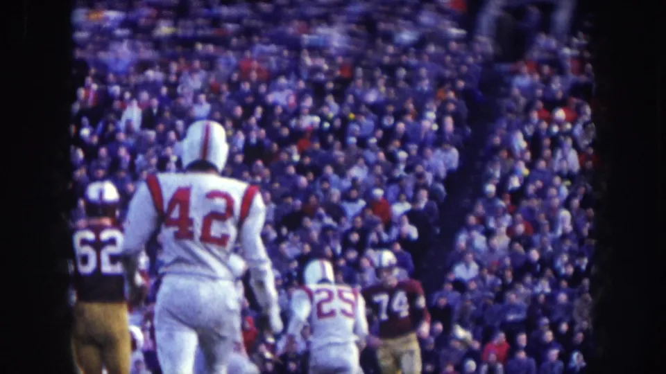 Sideline view at a 1957 Nebraska football game: players nos. 42, 25, 62, and 74 in white jerseys alongside dark-uniformed opponents, packed stands behind.