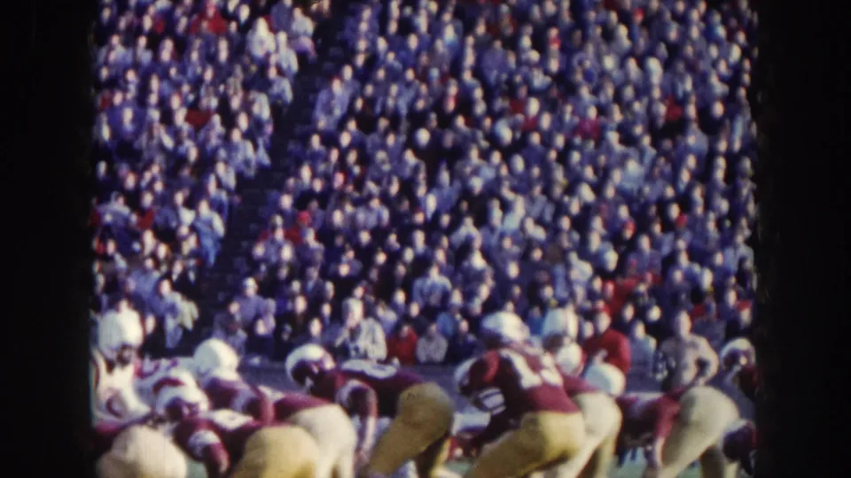 Close sideline view at a 1957 Nebraska football game: scrimmage pile of helmeted players in mixed uniforms, dense crowd filling the stands above.