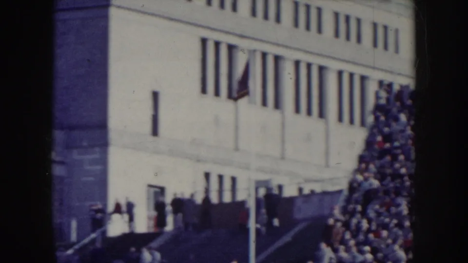 Mid-century institutional building with a flag, crowd gathered in foreground; may show a Nebraska university or civic venue circa 1957.