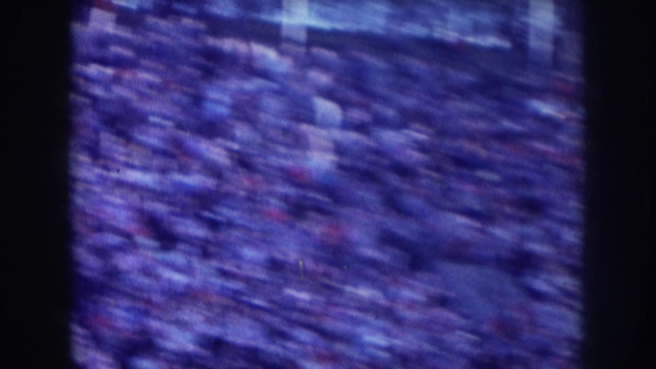 Tightly packed crowd filling the entire frame, blue-tinted grainy 8mm, small red and white clothing accents throughout.