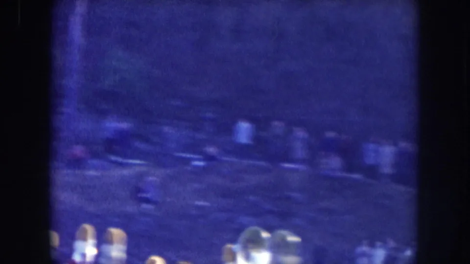Underexposed 8mm frame from a Nebraska parade, circa 1957; faint crowd silhouettes and small bright points of light visible at the lower edge only.