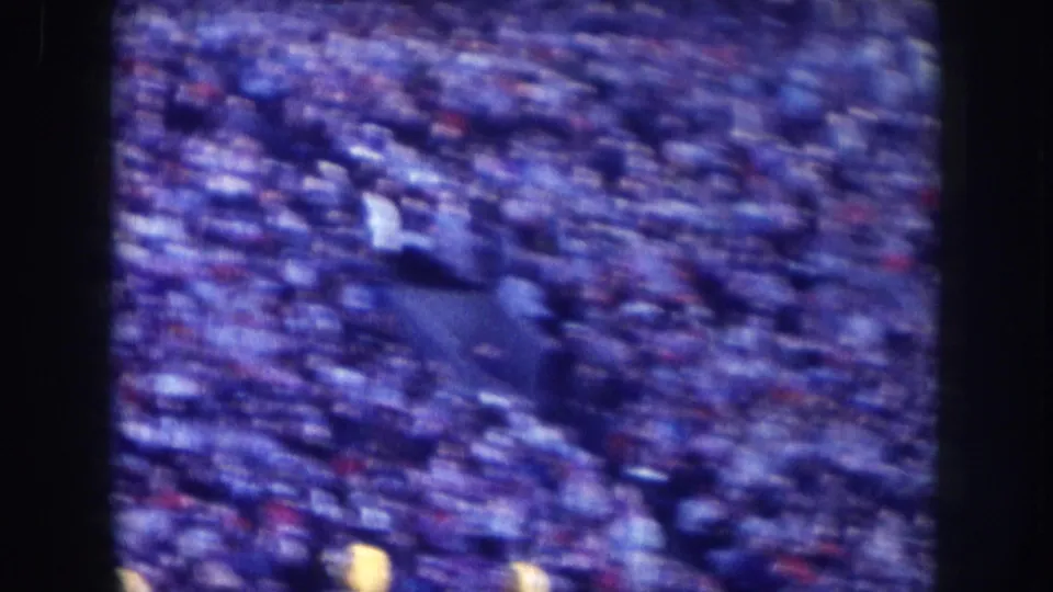 Blue-cast 8mm frame of a large parade crowd in Nebraska, circa 1957; masses of spectators with scattered red and white clothing accents, no identifiable individuals.