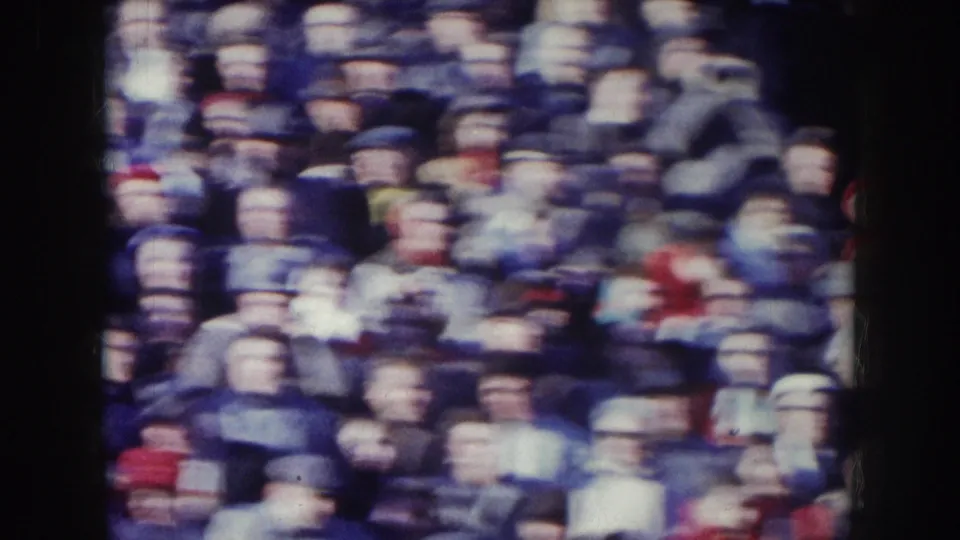 Closer 8mm framing of 1950s stadium spectators showing individual figures in hats and coats, consistent with a Nebraska football crowd circa 1957.