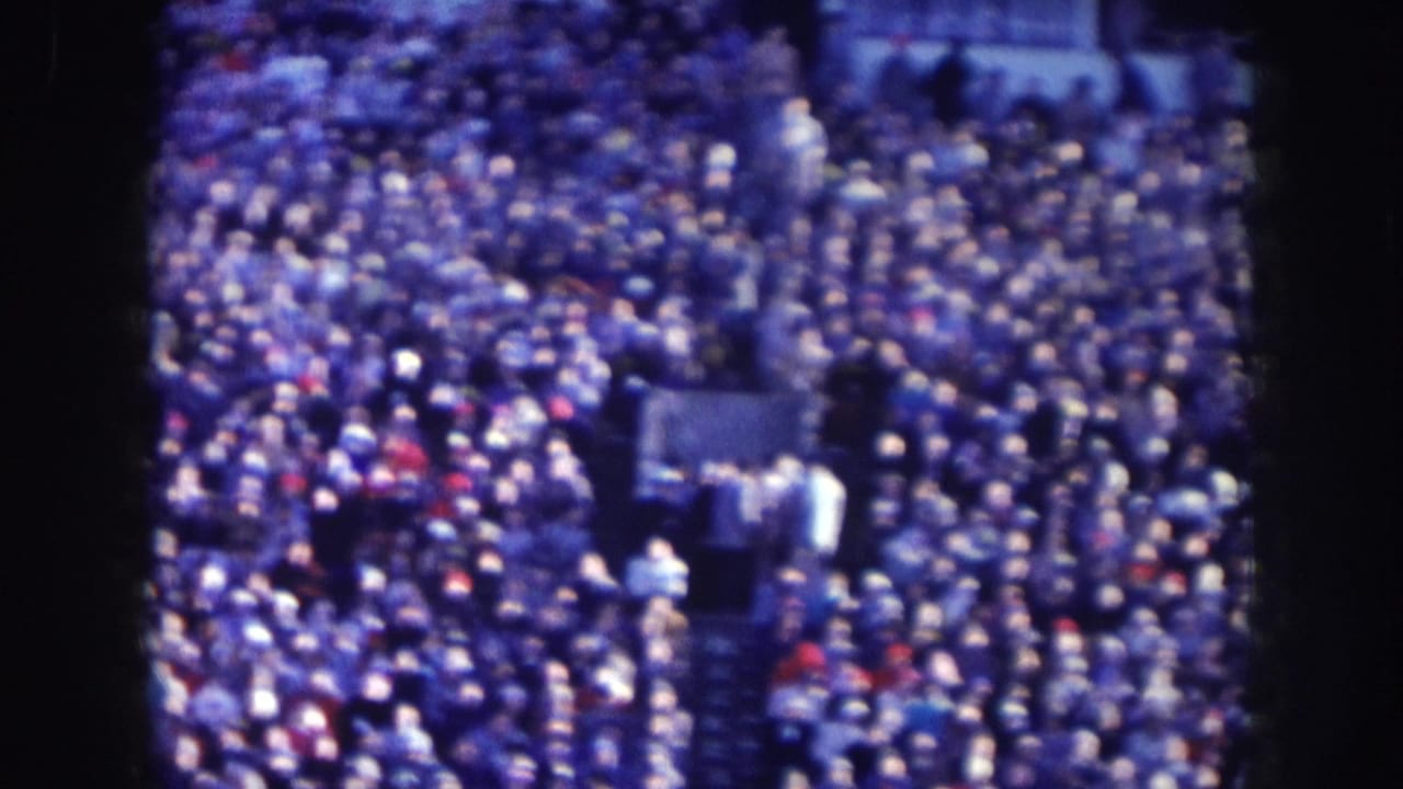 Elevated view of a dense crowd at an outdoor public gathering, 8mm film with blue tint and heavy grain.