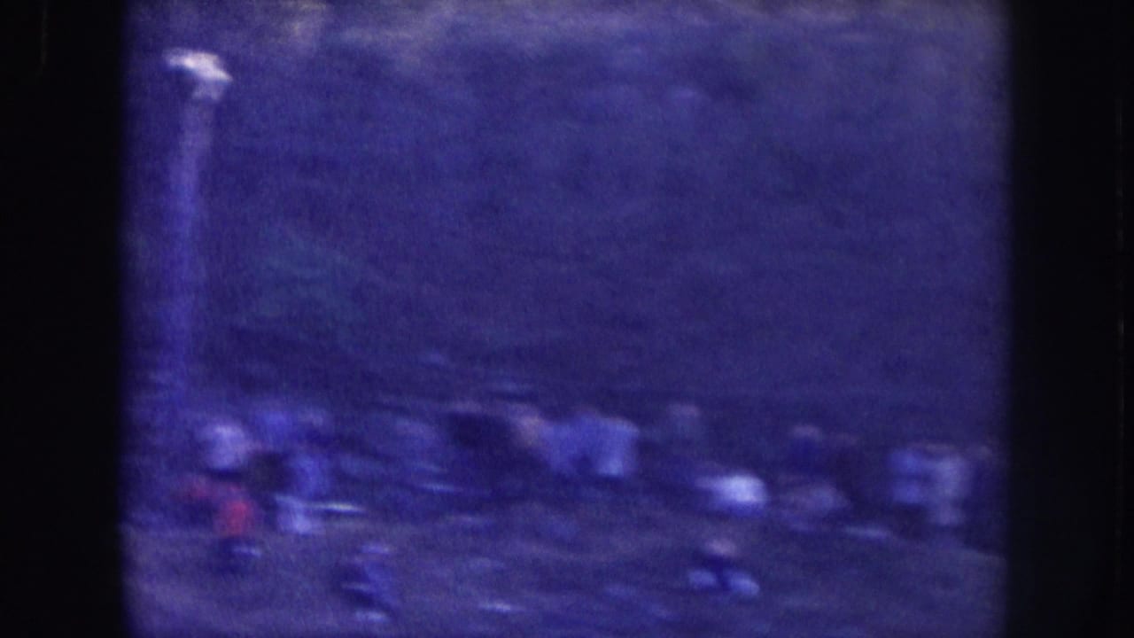 Several blurred figures outdoors, filmed on 8mm circa 1957 in Nebraska; deep blue-purple color shift and heavy grain render detail indistinct.