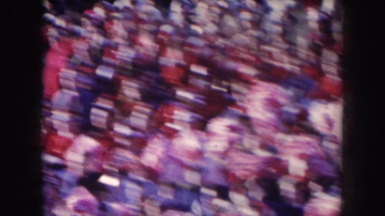 Motion-blurred 8mm wide shot of a packed stadium crowd wearing red and white, filmed from the stands at a 1957 Nebraska football game.