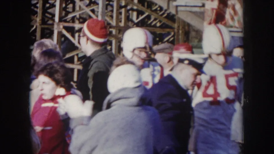 8mm close-up at sideline level showing a football player in white helmet and number 45 jersey among fans in winter coats, wooden stadium scaffolding visible, circa 1957 Nebraska.
