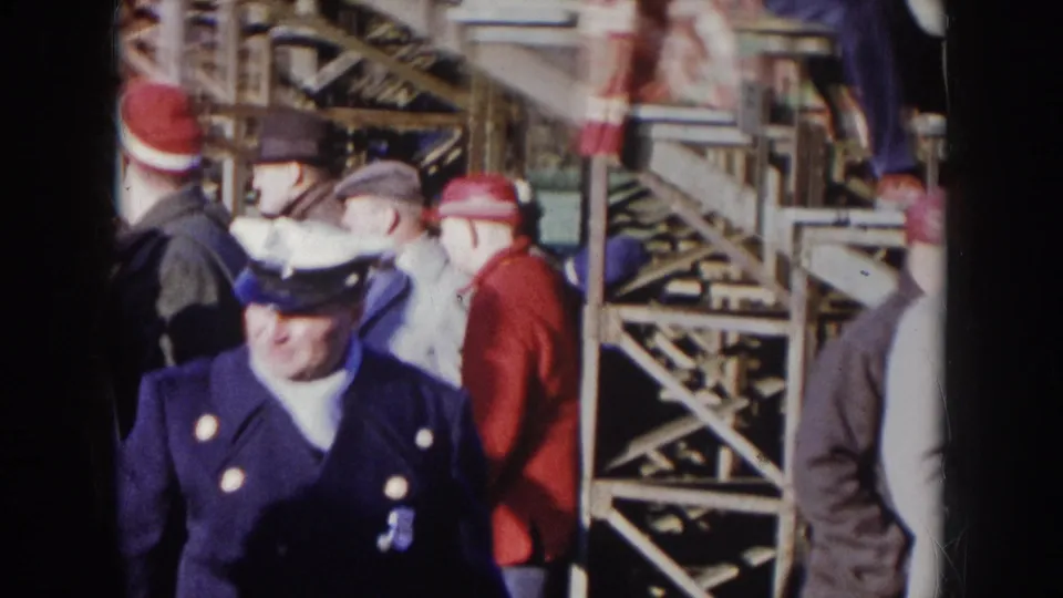 8mm ground-level shot of a uniformed stadium security officer in dark blue with brass buttons, surrounded by fans in red winter clothing, circa 1957 Nebraska football game.