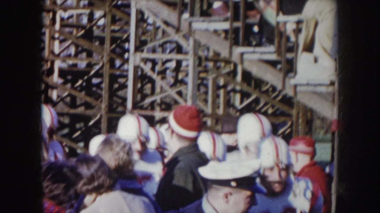 1957 Nebraska 8mm — football players in white helmets with red stripes push through spectators beside wooden scaffolding bleachers.