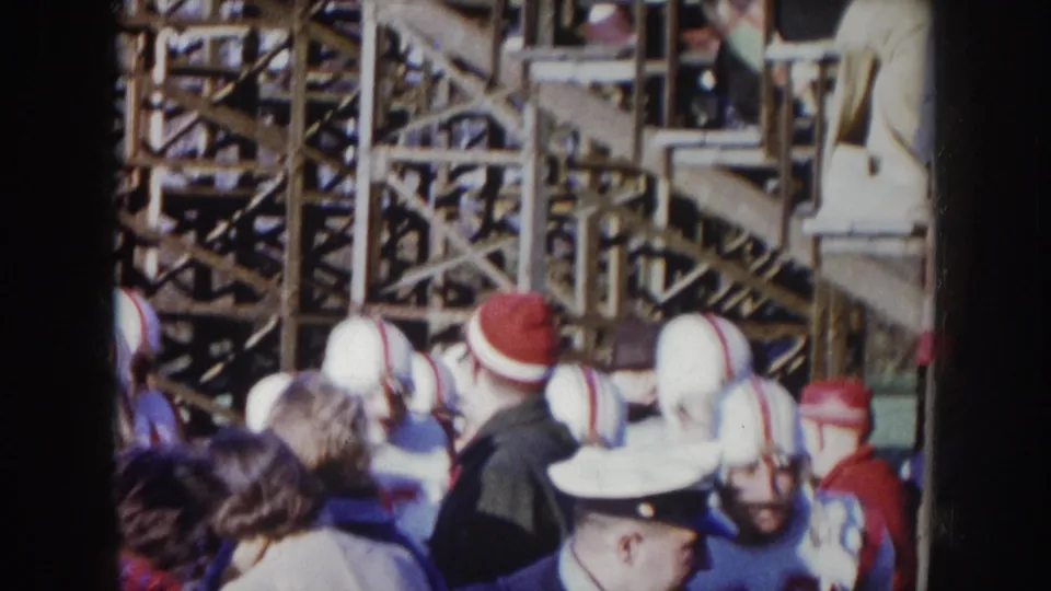 1957 Nebraska 8mm — football players in white helmets with red stripes push through spectators beside wooden scaffolding bleachers.