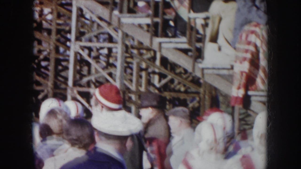 Football players in white uniforms with red-striped helmets amid winter-dressed spectators, wooden stands in background.