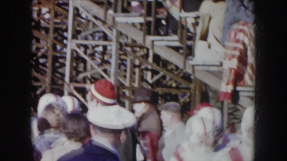 1957 Nebraska 8mm — players in white helmets and numbered jerseys moving through a crowd beside wooden bleacher scaffolding.