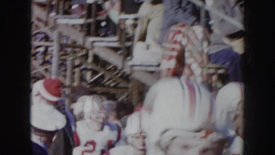 1957 Nebraska 8mm — close sideline view of players in white uniforms with red-striped helmets amid a densely packed winter crowd.