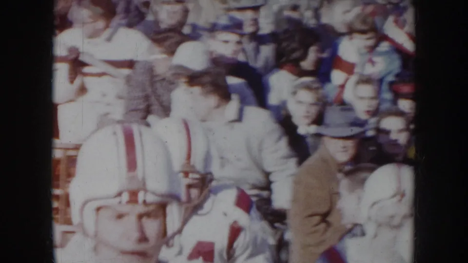 1957 Nebraska 8mm — player in white helmet and jersey number 4 surrounded by spectators in winter hats and coats at wooden bleachers.