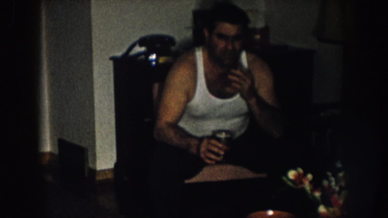 Man in white undershirt seated in a dimly lit home interior, circa 1957 Nebraska; dark wood dresser and artificial flower arrangement visible behind him.