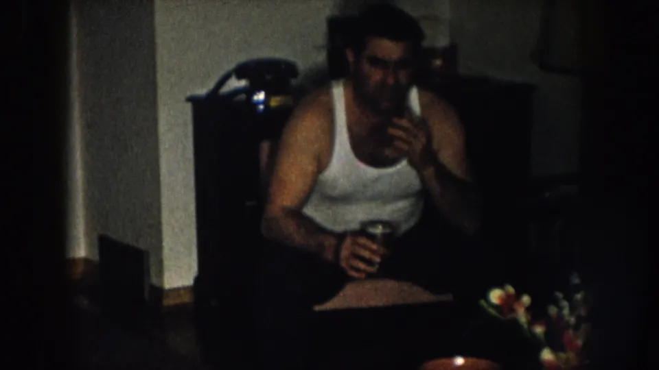 Man in white undershirt seated in a dimly lit home interior, circa 1957 Nebraska; dark wood dresser and artificial flower arrangement visible behind him.