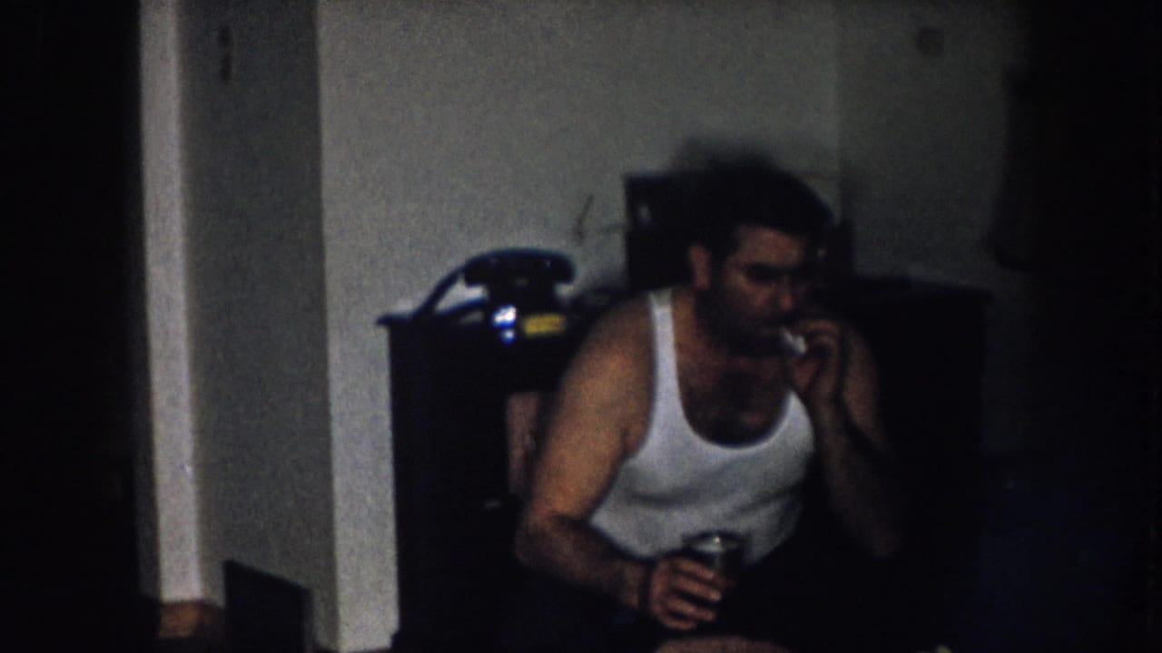 Man in white tank top looks down while holding a beverage can; dark furniture with what may be a rotary telephone visible in background at upper right.