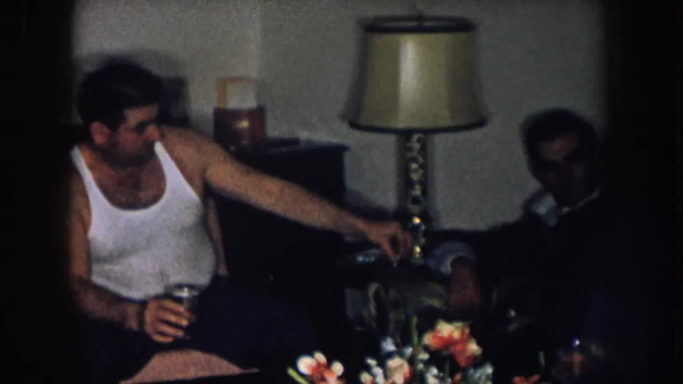 Man in white undershirt in 1950s home interior beside a mid-century table lamp; second person partially visible at right edge, artificial flowers in foreground.