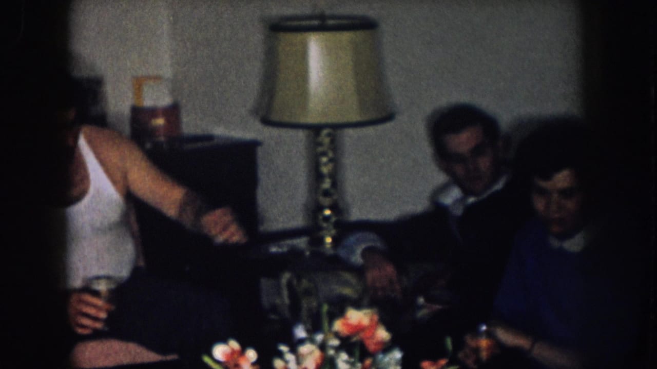 Man in white tank top holding a glass reaches across the frame toward a couple seated on a sofa; table lamp and cut flowers visible in background.