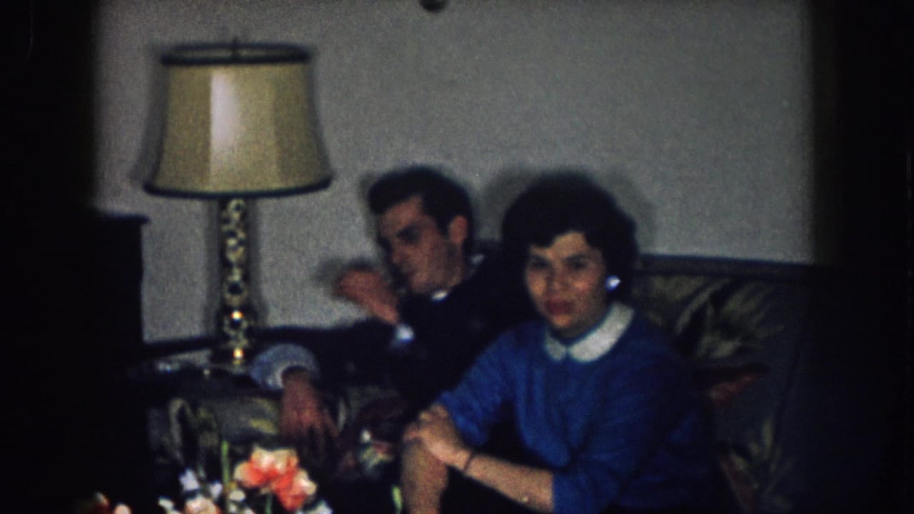 Man in dark jacket and woman in blue sweater with white collar seated on a dark sofa, table lamp behind them, cut flower arrangement in foreground.