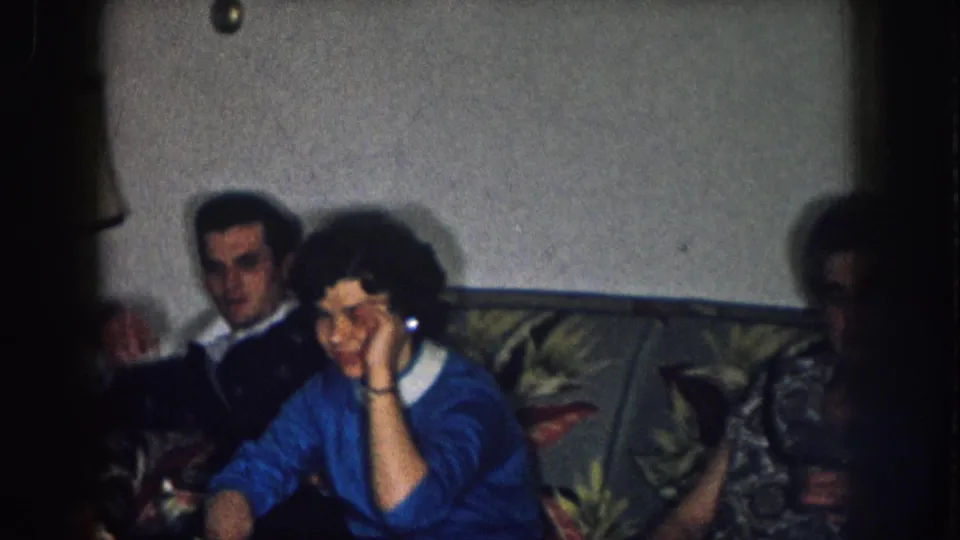 1957 Nebraska home movie: man in dark jacket and woman in blue sweater on floral-upholstered sofa, woman gesturing near her face, partial third figure behind.
