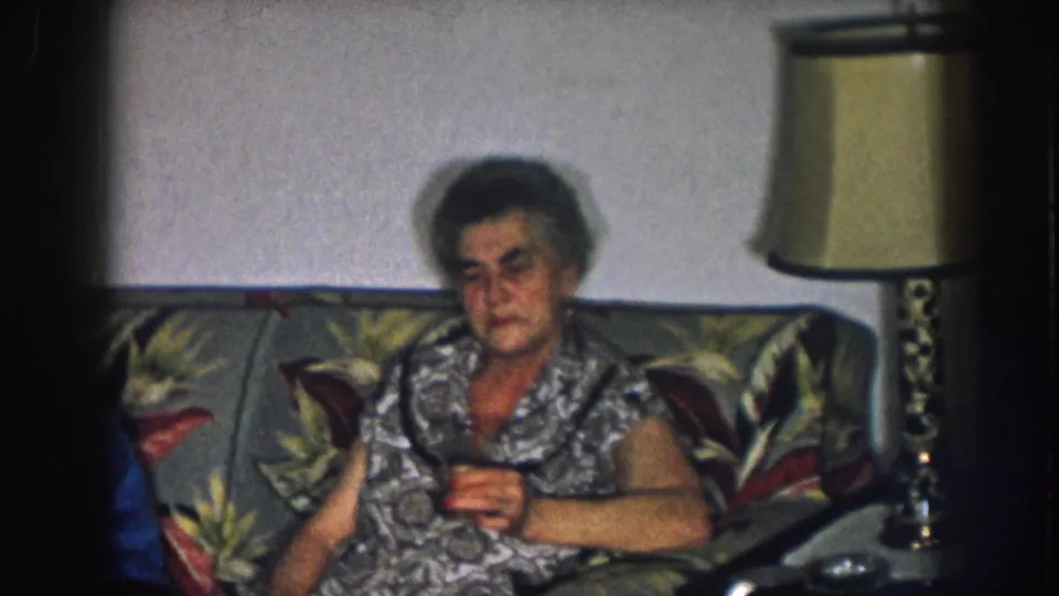 1957 Nebraska home movie: older woman in printed housedress seated alone on a tropical-print sofa, table lamp behind her in a domestic living room.