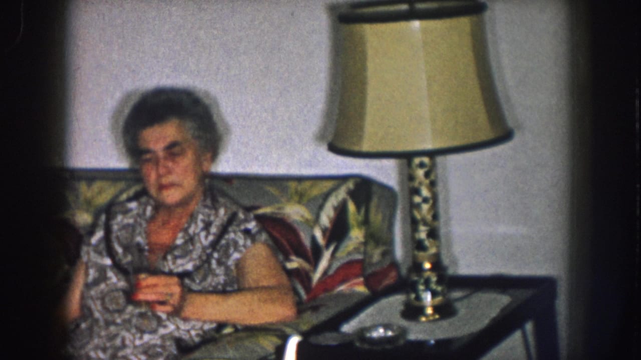 Older woman in a patterned house dress seated on a floral sofa holding a small glass; brass lamp on side table; Nebraska living room, circa 1957.