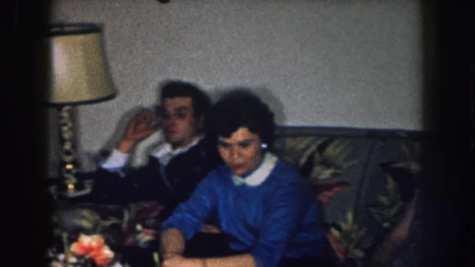 Woman in blue sweater with white collar and a second person in the foreground; floral couch and brass lamp behind; cut flowers at lower frame; Nebraska home, circa 1957.