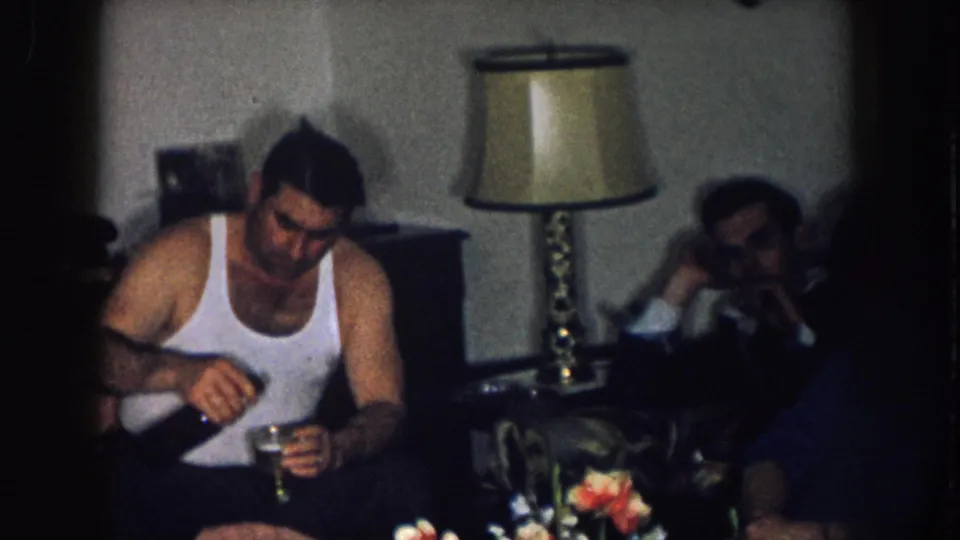 Young man in a white undershirt holding a small glass in a living room; brass lamp and cut flowers visible at right; Nebraska home, circa 1957.