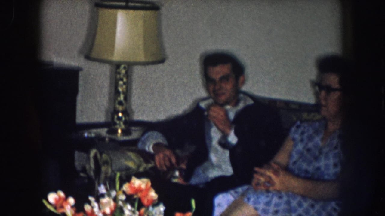 1950s home movie still: man in dark jacket and woman in blue patterned dress with cat-eye glasses on a sofa; brass lamp and floral arrangement visible, Nebraska, circa 1957.