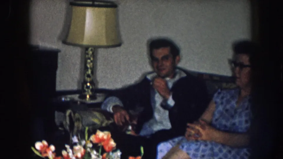 1950s home movie still: man in dark jacket and woman in blue patterned dress with cat-eye glasses on a sofa; brass lamp and floral arrangement visible, Nebraska, circa 1957.