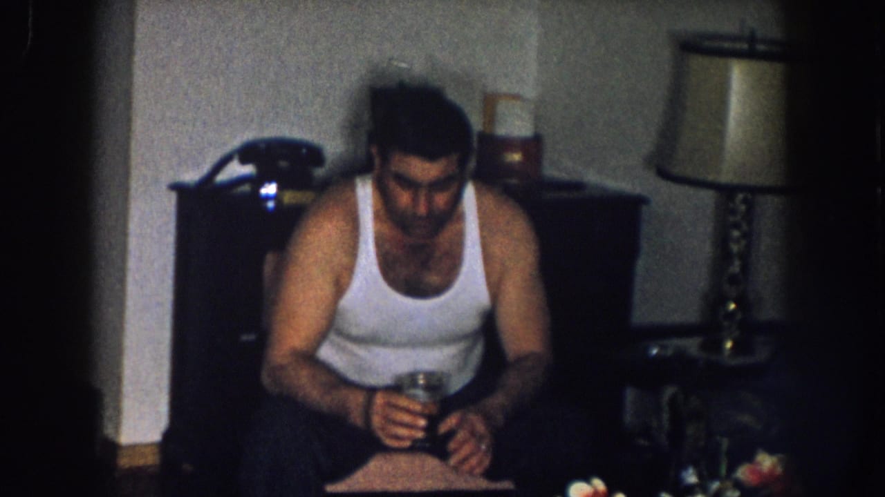 Man in white sleeveless undershirt seated and looking down, holding a glass; brass table lamp and orange flowers visible in background.