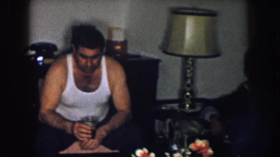 8mm home movie frame: man in a white sleeveless undershirt seated and looking down, brass table lamp visible in center, dark object resembling a telephone on background furniture, Nebraska, circa 1957.