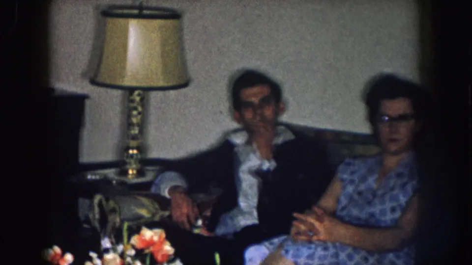 Late-1950s 8mm home movie: man in dark jacket and woman in blue patterned dress with cat-eye glasses seated on a sofa beneath a brass table lamp, Nebraska, circa 1957.
